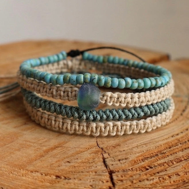 A set of three woven wrap bracelets with small beads in light blue, dark gray, and beige, featuring a central iridescent blue bead, displayed on a rustic wooden slice.