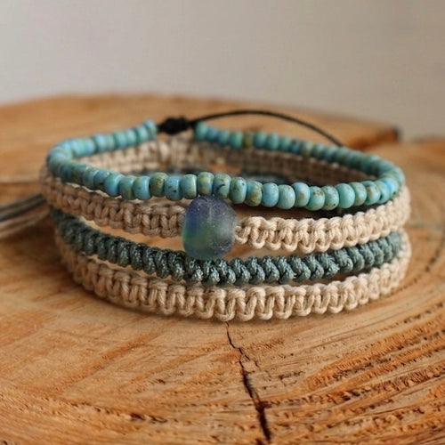 A set of three woven wrap bracelets with small beads in light blue, dark gray, and beige, featuring a central iridescent blue bead, displayed on a rustic wooden slice.