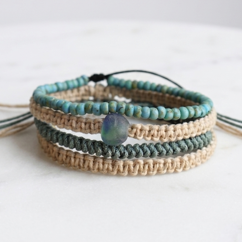 A stack of four bohemian-style beaded and woven bracelets on a white marble surface. The bracelets feature turquoise beads, natural tan woven cords, and a central seaglass stone.