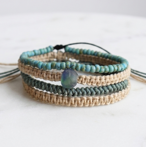 A stack of four bohemian-style beaded and woven bracelets on a white marble surface. The bracelets feature turquoise beads, natural tan woven cords, and a central seaglass stone.