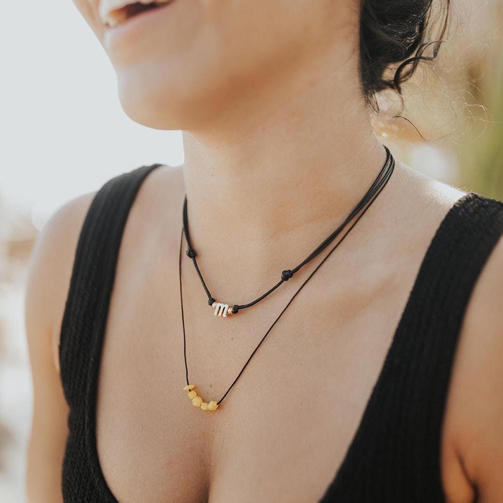 Personalized initial letter charm necklace on a black waxed cord, worn layered on a woman's neck.