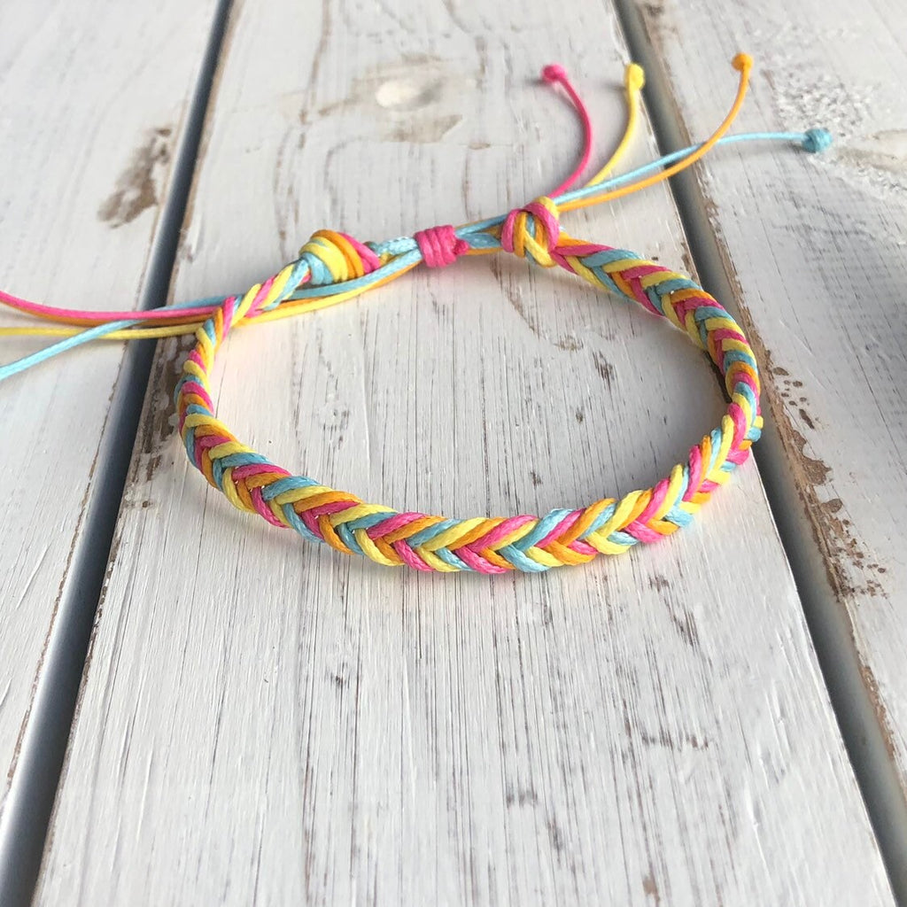 Colorful braided friendship bracelet in rainbow pastel tones, displayed on a rustic white wooden surface with adjustable string closure.