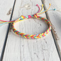Colorful braided friendship bracelet in rainbow pastel tones, displayed on a rustic white wooden surface with adjustable string closure.