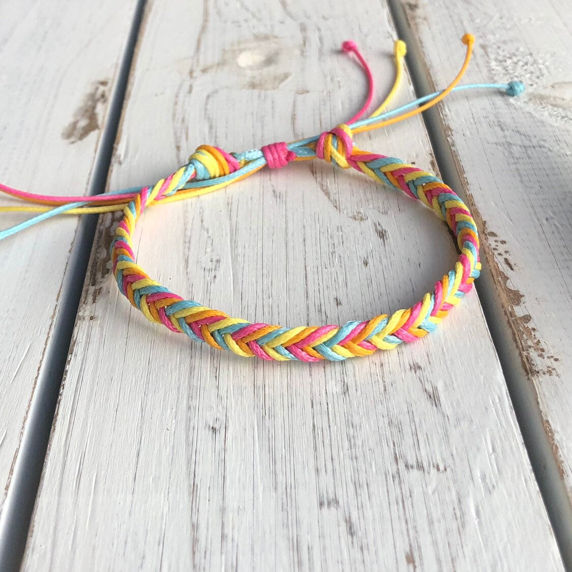 Colorful braided friendship bracelet in rainbow pastel tones, displayed on a rustic white wooden surface with adjustable string closure.