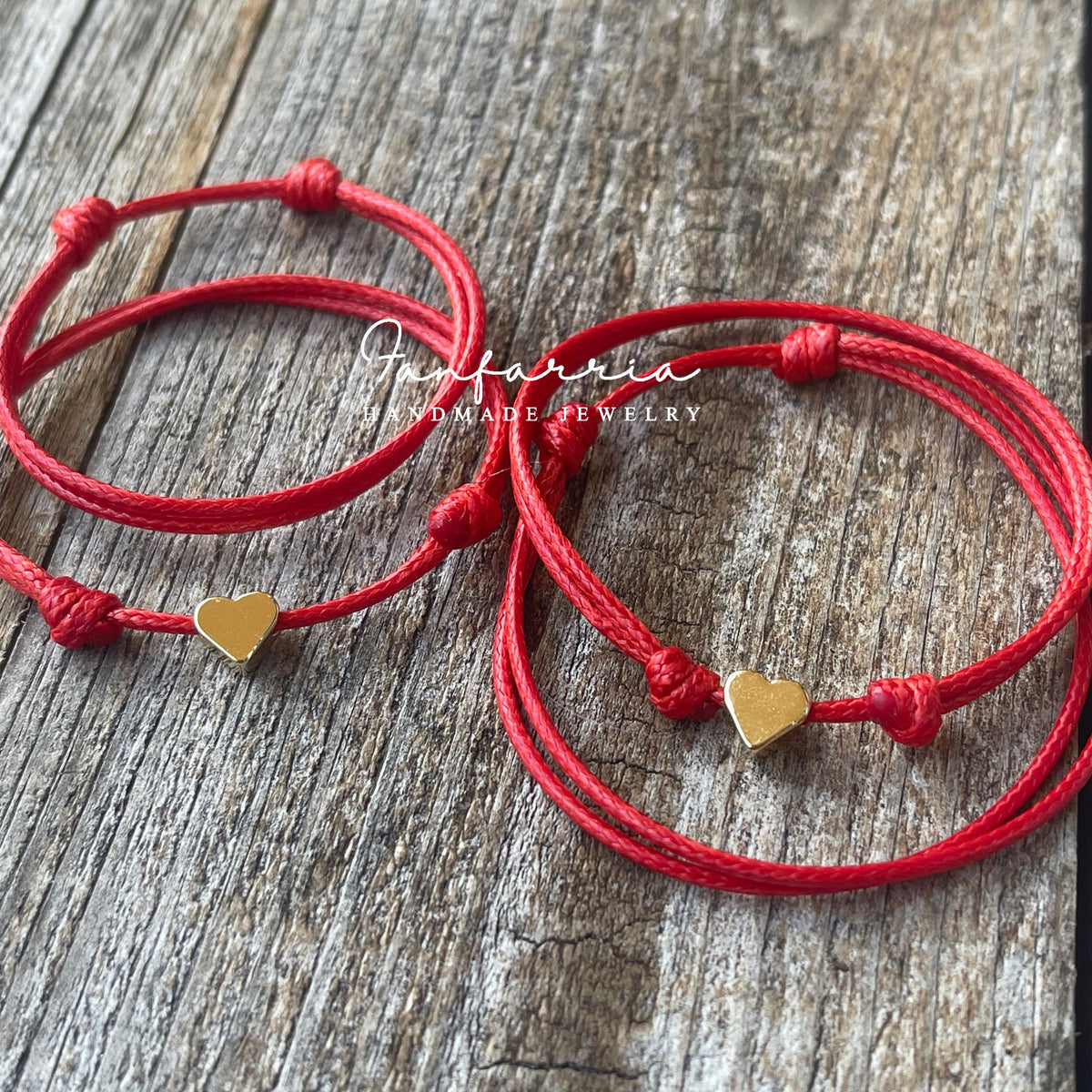 May include: Red string bracelets with gold heart charms. The bracelets are tied in knots and are on a wooden surface.