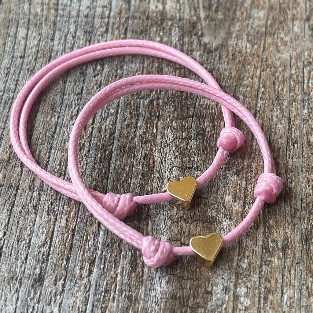 Two pink friendship bracelets with gold heart charms. The bracelets are tied together with a knot.