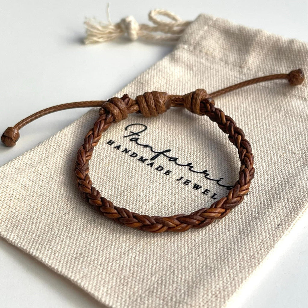 A brown leather braided bracelet with a knot closure. The bracelet is displayed on a white fabric bag with the text &#39;Tanparis Handmade Jewel&#39; printed on it.