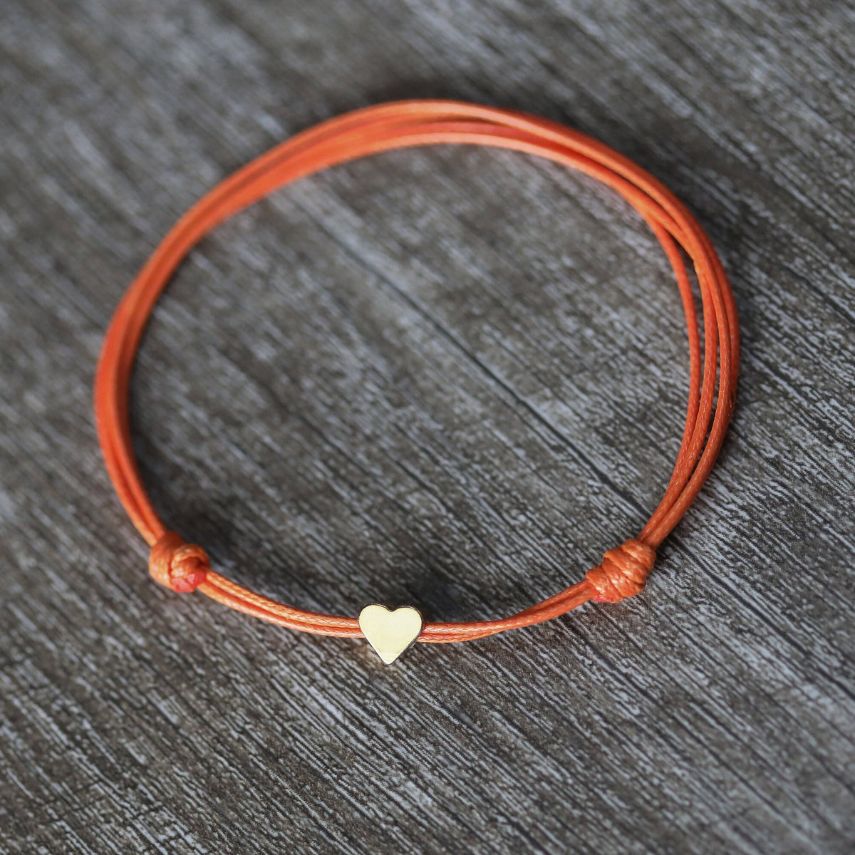 A simple orange string anklet bracelet with a small gold heart charm.