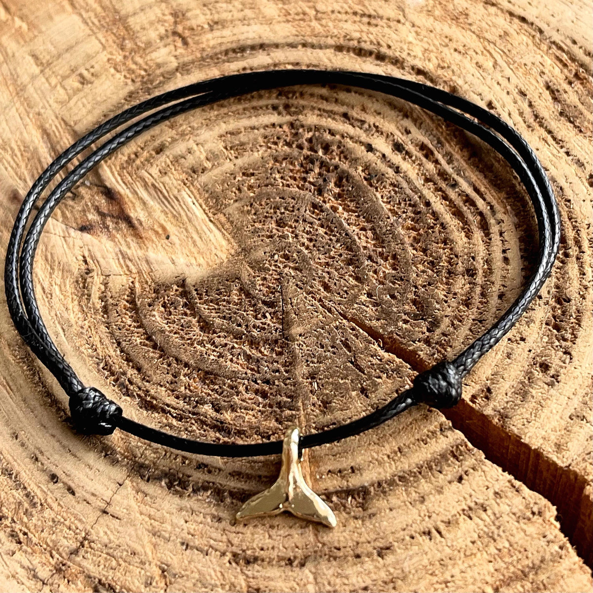 Black cord anklet with a gold plated whale tail charm. The anklet is knotted and rests on a wooden surface.