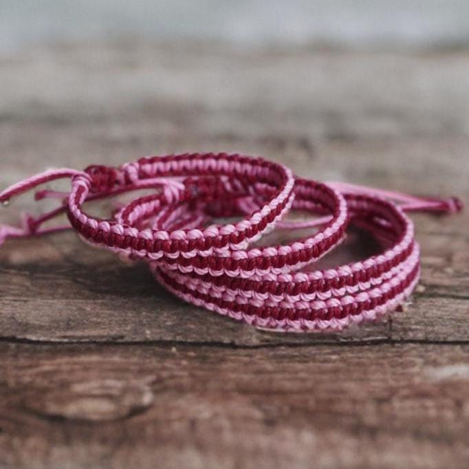 Handmade pink and burgundy macrame bracelets stacked on a rustic wooden surface, adjustable woven cord design, boho beach style friendship bracelets.