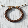 Handmade braided brown leather bracelet with adjustable cord on a rustic white wooden surface.