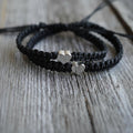 Two black macrame bracelets with silver heart charms. The bracelets are knotted and have a simple, elegant design. The background is a weathered wooden surface.