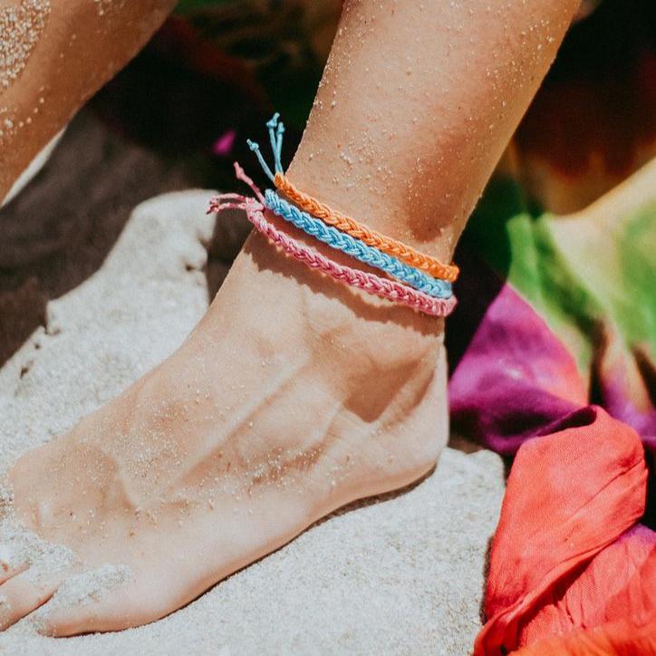 Braided orange, turquoise, and pink cord hemp anklets stacked on a sandy foot at the beach.