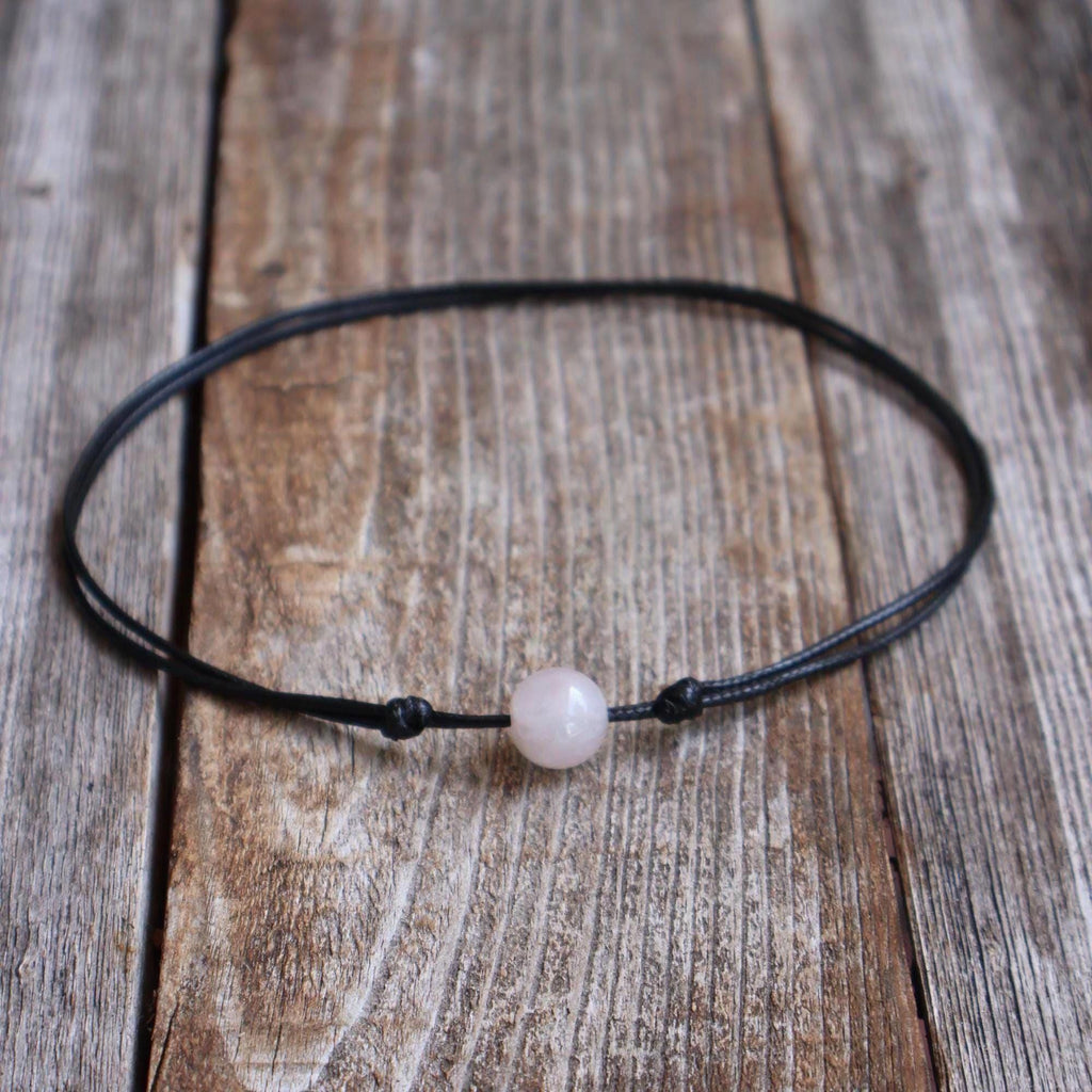 Rose quartz stone bead choker on black waxed cord with adjustable slide-knot closure, laid on a rustic wooden background.