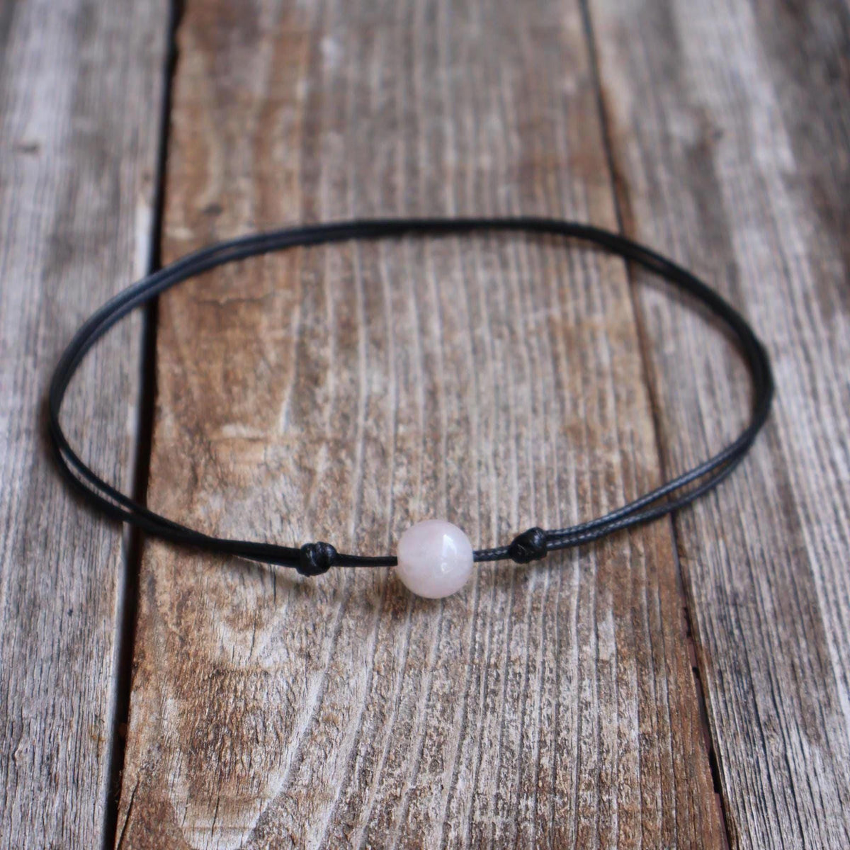 Rose quartz stone bead choker on black waxed cord with adjustable slide-knot closure, laid on a rustic wooden background.