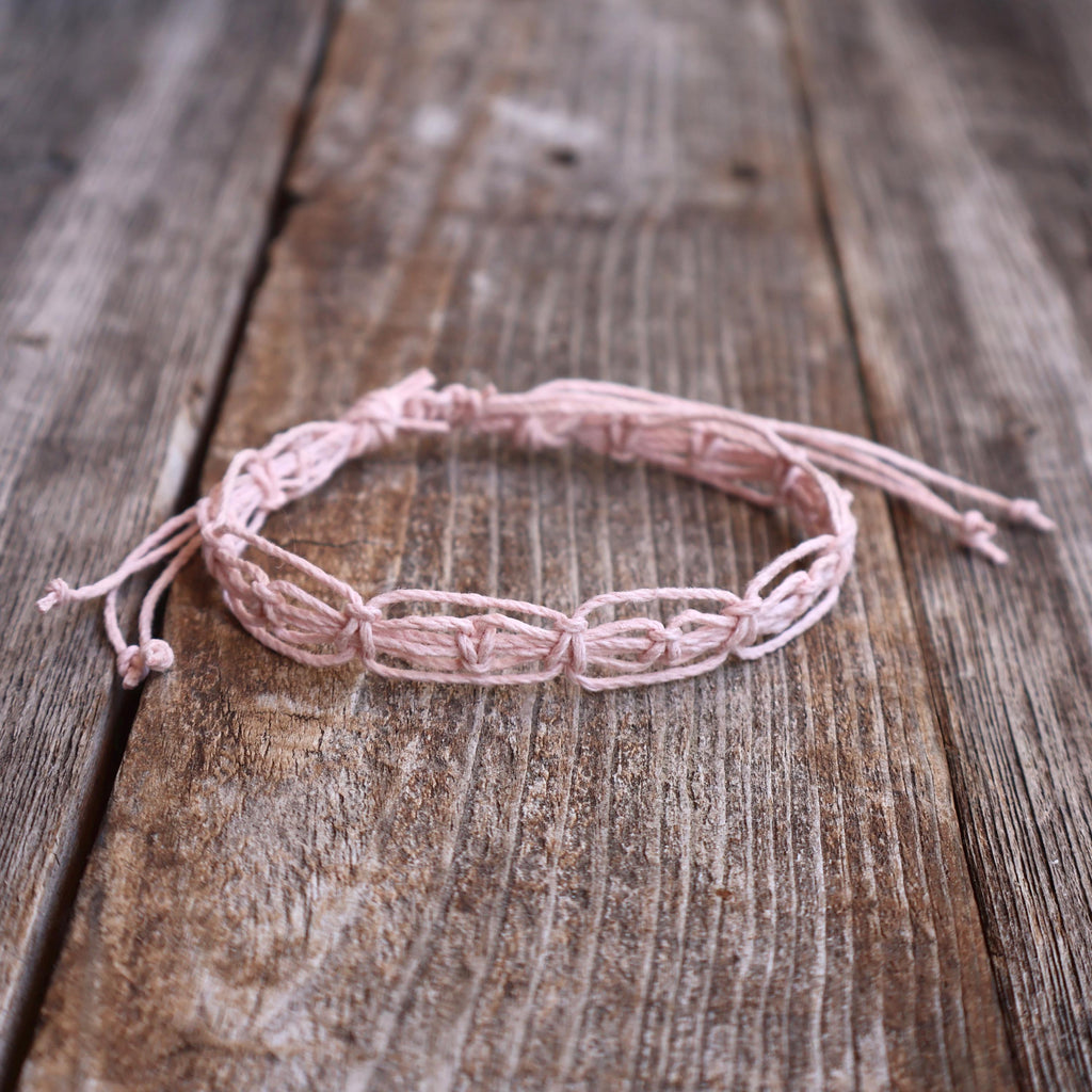 Blush pink macrame woven bracelet or anklet with adjustable slide-knot closure, laid on a rustic wooden background