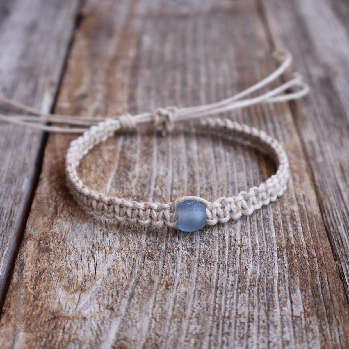 Natural macrame bracelet with blue sea glass bead accent, adjustable cord closure, on rustic wood background