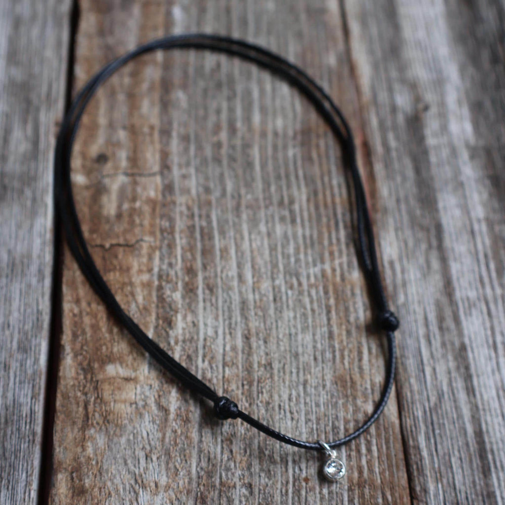 Swarovski birthstone crystal charm pendant on a black waxed cord necklace with adjustable slide-knot closure, laid on a rustic wooden background