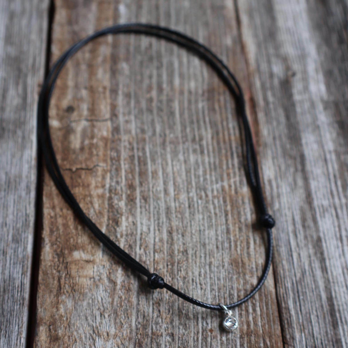 Swarovski birthstone crystal charm pendant on a black waxed cord necklace with adjustable slide-knot closure, laid on a rustic wooden background