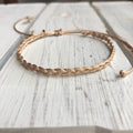 May include: A braided, adjustable bracelet in a light tan color. The bracelet features a woven design and a sliding knot closure. The background is a white wooden surface.