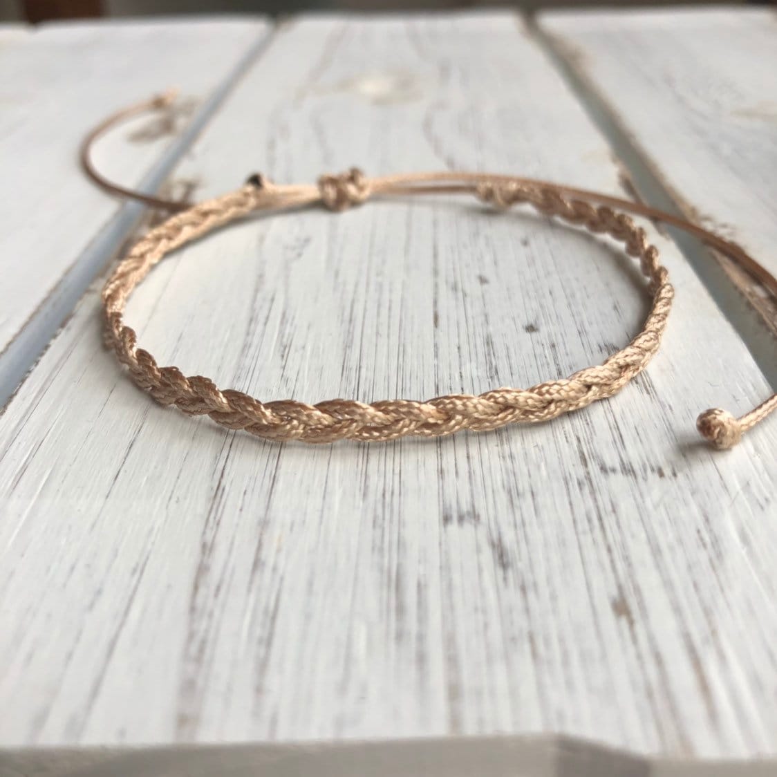 May include: A braided, adjustable bracelet in a light tan color. The bracelet features a woven design and a sliding knot closure. The background is a white wooden surface.