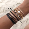 Boho bracelet stack with freshwater pearl, gold beads, and braided black, white, and blush cord bracelets worn on wrist at the beach