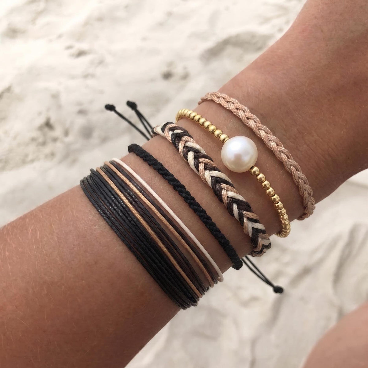Boho bracelet stack with freshwater pearl, gold beads, and braided black, white, and blush cord bracelets worn on wrist at the beach