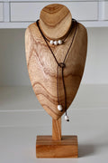 May include: A lariat-style necklace featuring a dark brown leather cord adorned with five white, smooth, oval-shaped pearls. Three pearls are positioned horizontally along the upper section, while two pearls hang at the ends of the cord.
