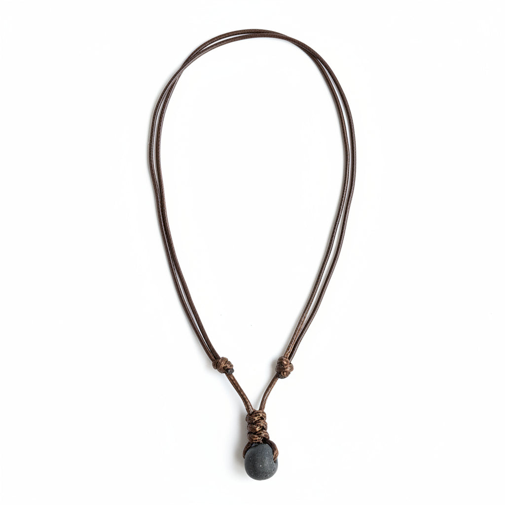 May include: A brown leather necklace with a dark gray stone pendant. The necklace is knotted and hangs against a weathered wooden background. The stone is smooth and round.