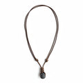 May include: A brown leather necklace with a dark gray stone pendant. The necklace is knotted and hangs against a weathered wooden background. The stone is smooth and round.