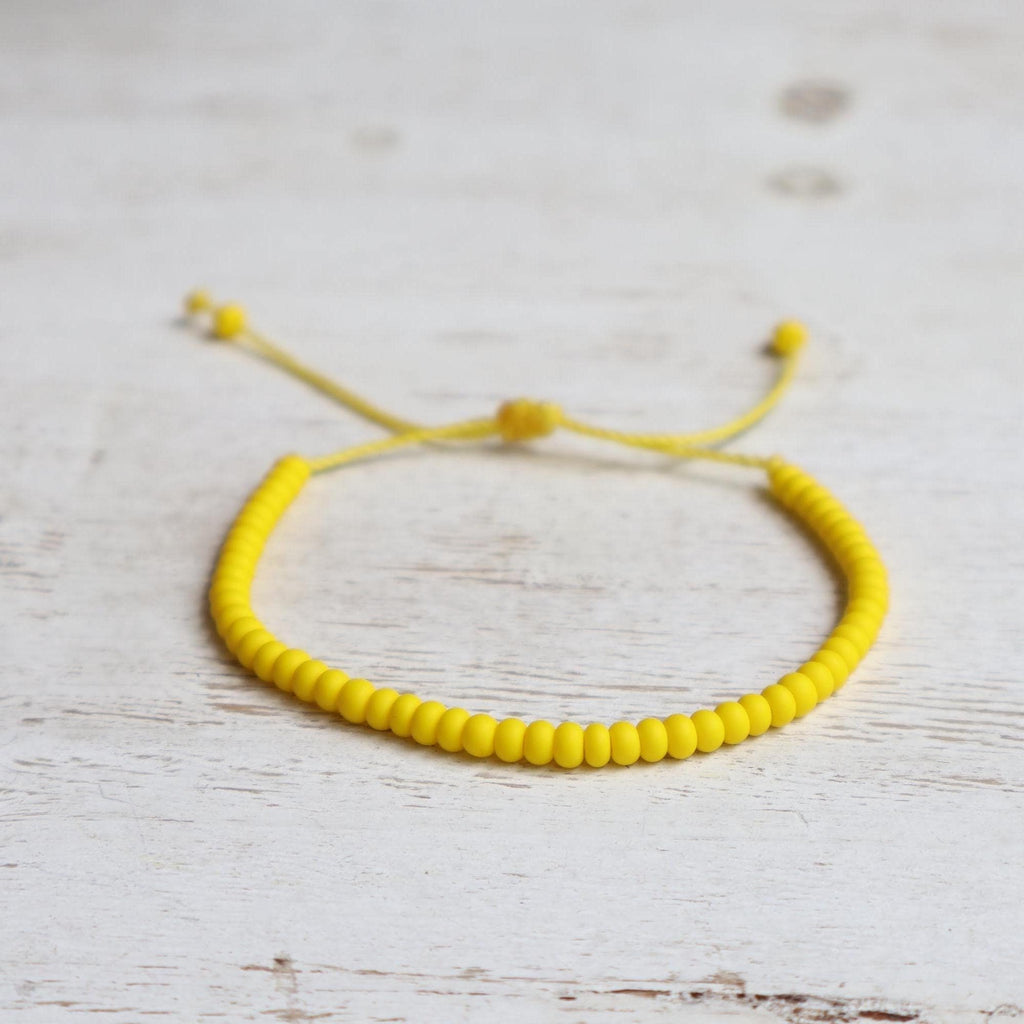 Minimalist yellow bracelet made with 6/0 Czech glass seed beads and an adjustable slide-knot cord closure, photographed on a rustic white wood background.