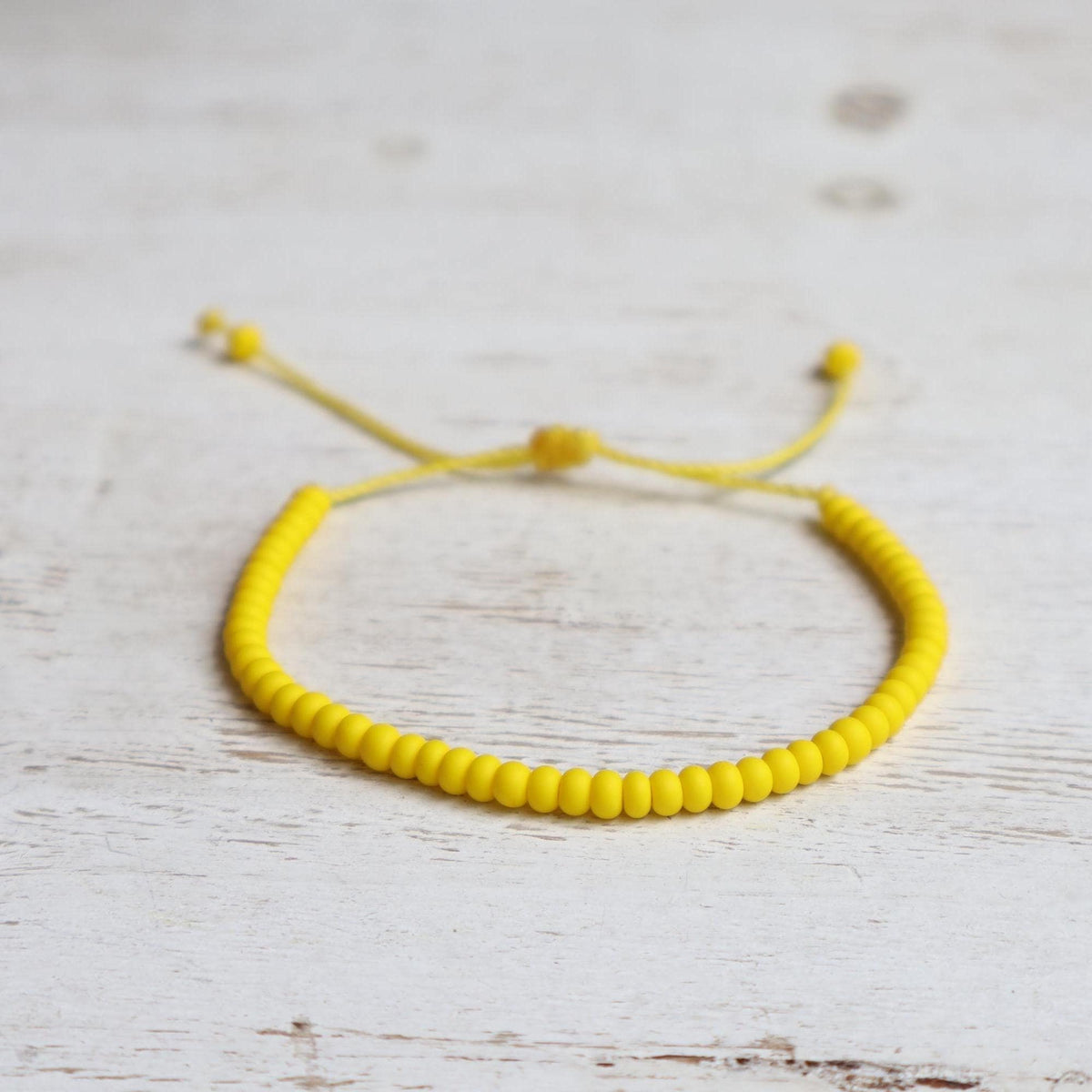 Minimalist yellow bracelet made with 6/0 Czech glass seed beads and an adjustable slide-knot cord closure, photographed on a rustic white wood background.