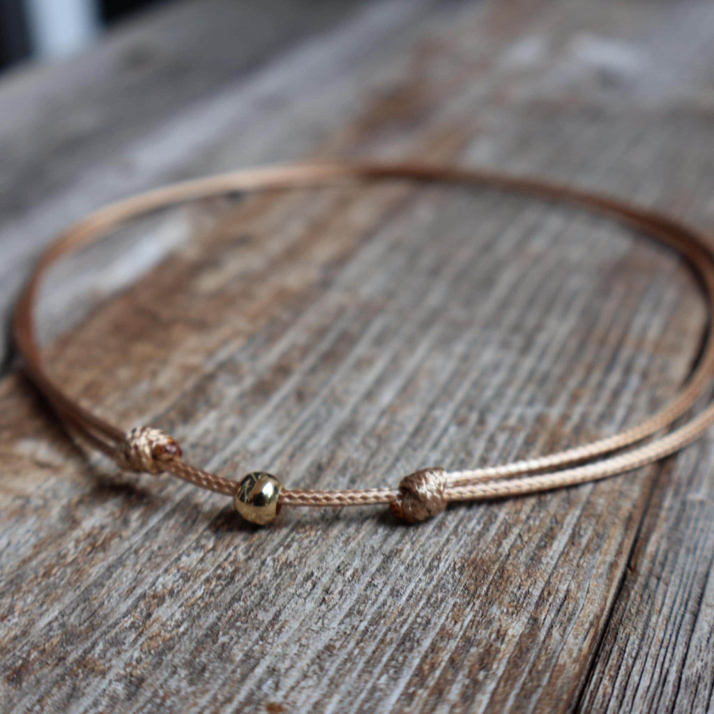 Minimalist tan waxed cord choker necklace with 6mm gold bead and adjustable knot closure, displayed on a wood background