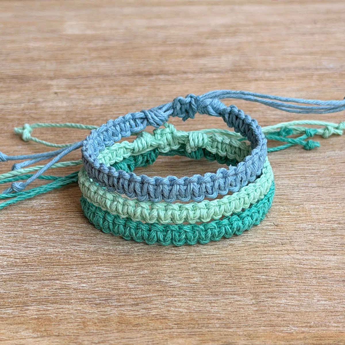 May include: A set of three stacked bracelets crafted from hemp cord in varying shades of blue and green. The bracelets feature a macrame knotted pattern and showcase light blue, mint, and teal.