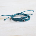 Personalized matching initial cord bracelets for long-distance couples, showcasing teal braided bands with P and E letter beads on a light wooden table.