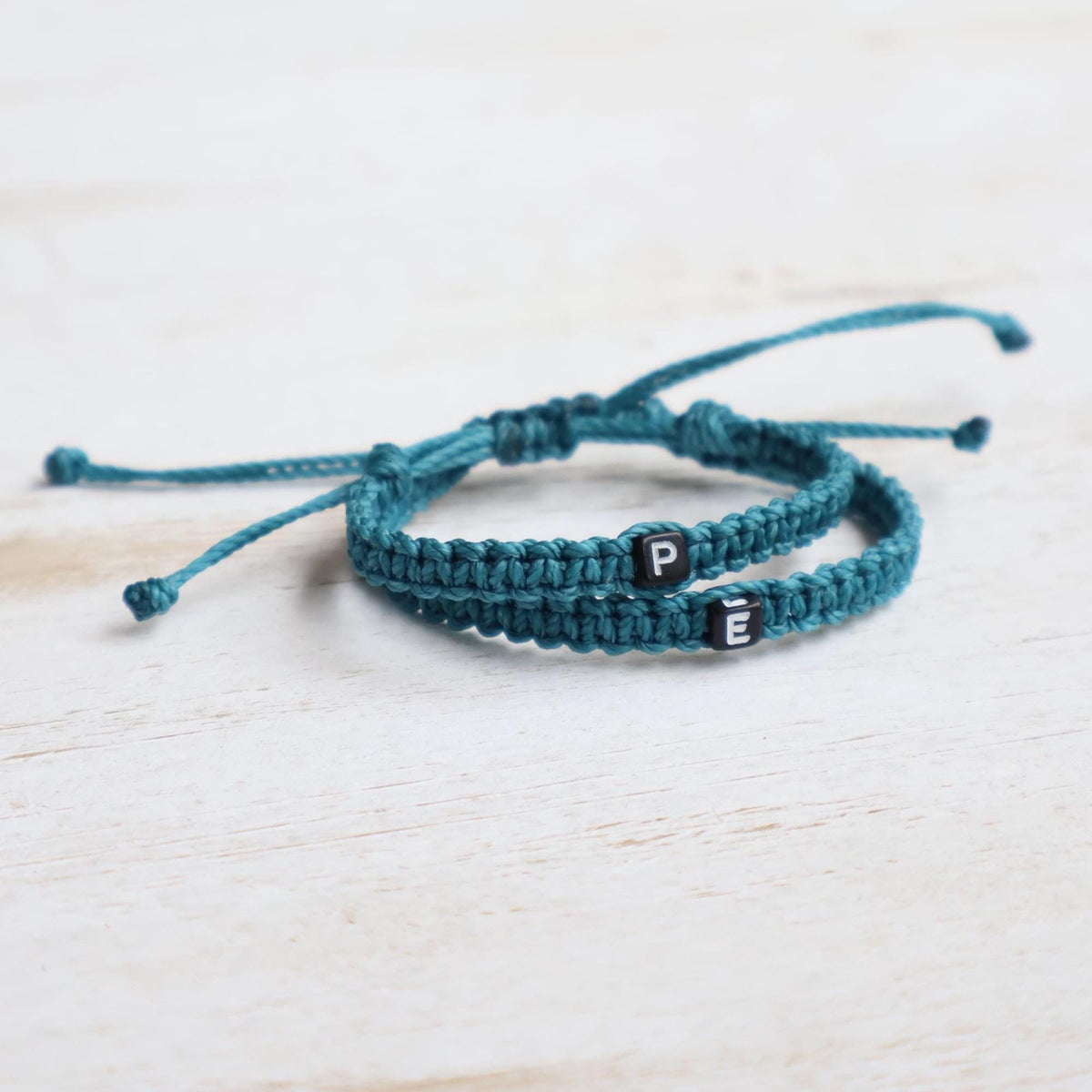 Personalized matching initial cord bracelets for long-distance couples, showcasing teal braided bands with P and E letter beads on a light wooden table.