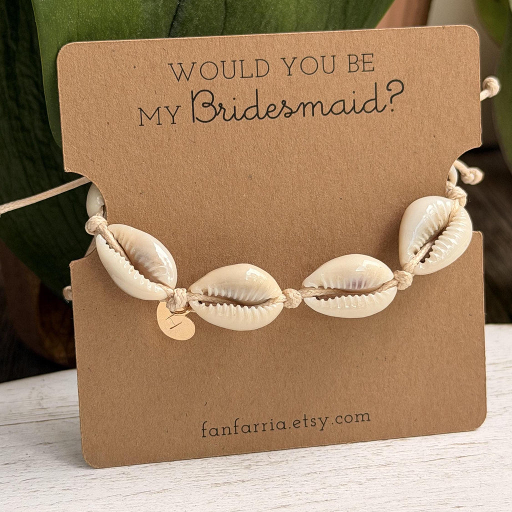 Cowrie shell bridesmaid bracelet displayed on a kraft paper 'Would You Be My Bridesmaid?' proposal card with a gold personalized initial charm, set against a green leaf background.