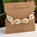 Cowrie shell bridesmaid bracelet displayed on a kraft paper 'Would You Be My Bridesmaid?' proposal card with a gold personalized initial charm, set against a green leaf background.