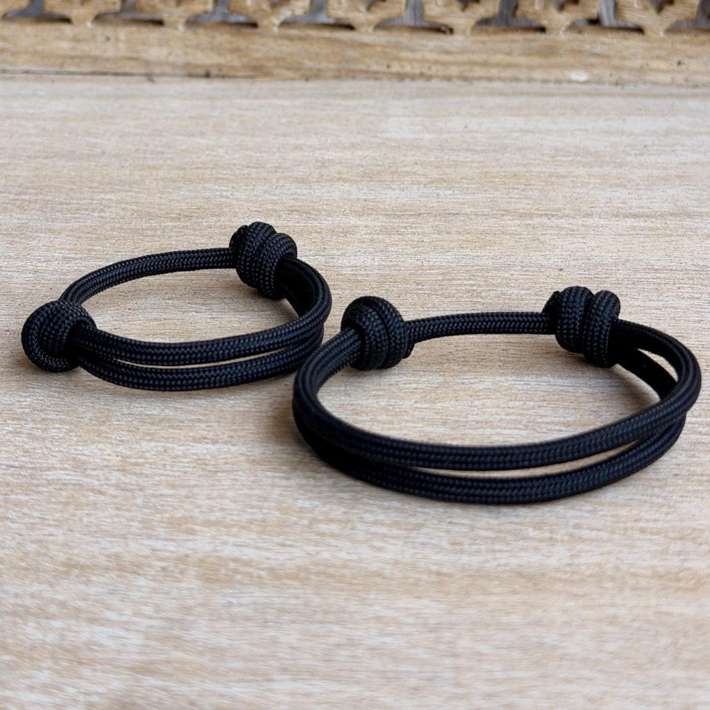 Matching black paracord bracelets for adult and child with adjustable sliding knots, minimalist unisex family bracelet set on a light wood background.