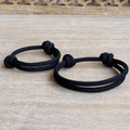 Matching black paracord bracelets for adult and child with adjustable sliding knots, minimalist unisex family bracelet set on a light wood background.