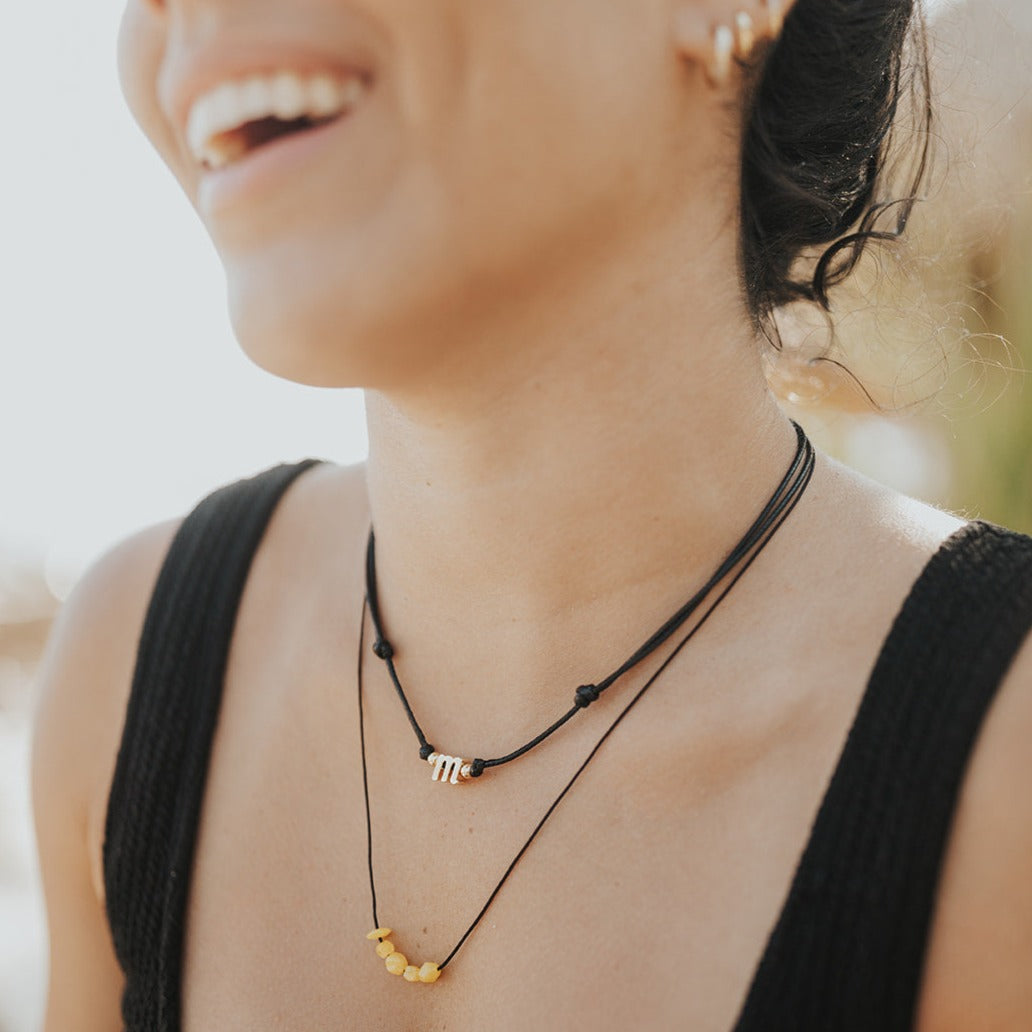 A person wearing a black top and showcasing two choker necklaces, one with a gold-plated initial and the other with yellow beads.